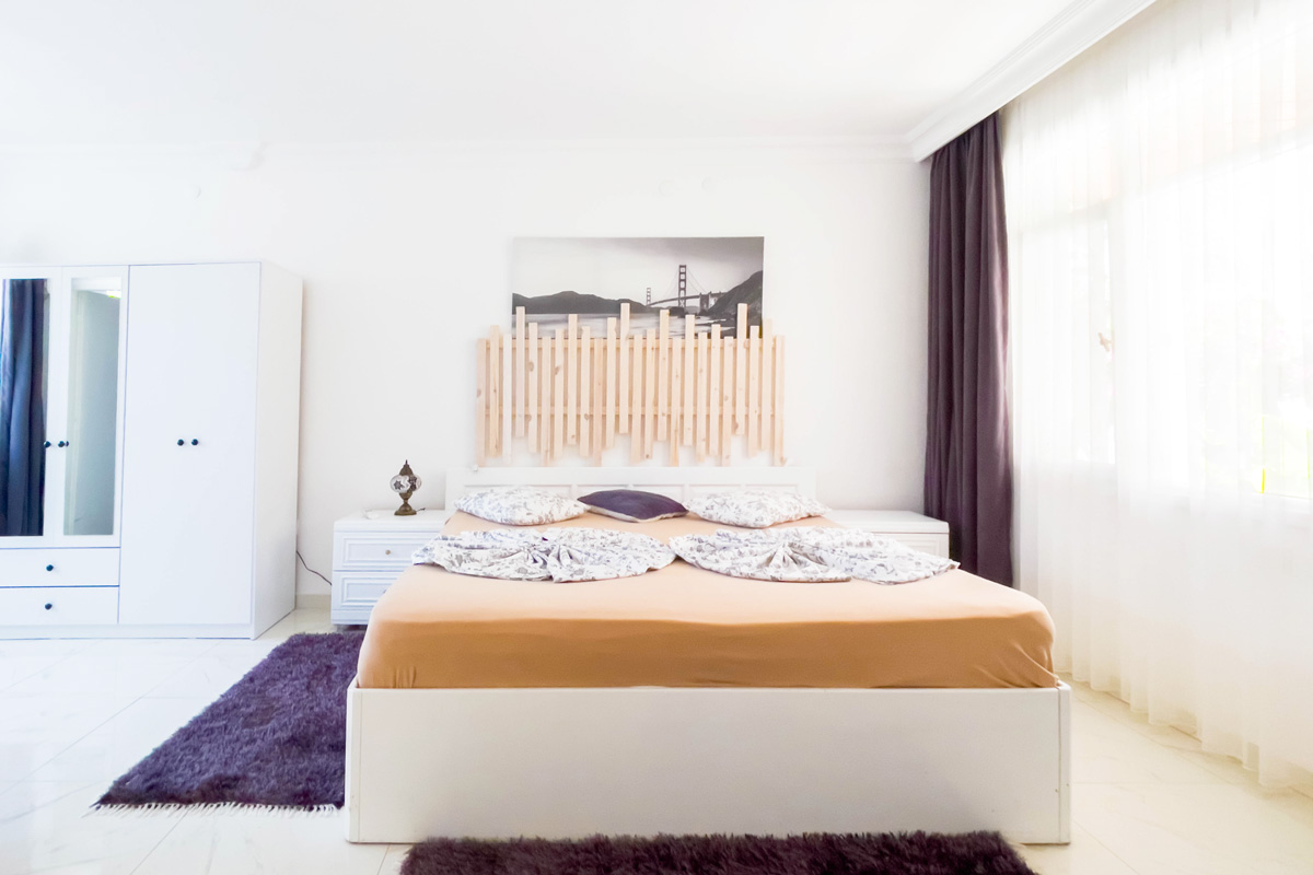 Bright and cozy bedroom featuring a large bed with a wooden headboard, soft neutral bedding, white furniture, and a large window with sheer curtains letting in natural light.