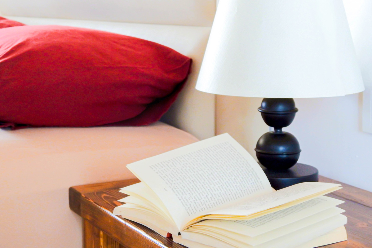 Cozy bedroom detail with an open book on a wooden bedside table, next to a black lamp with a white shade, and a bed with deep red pillows in the background.