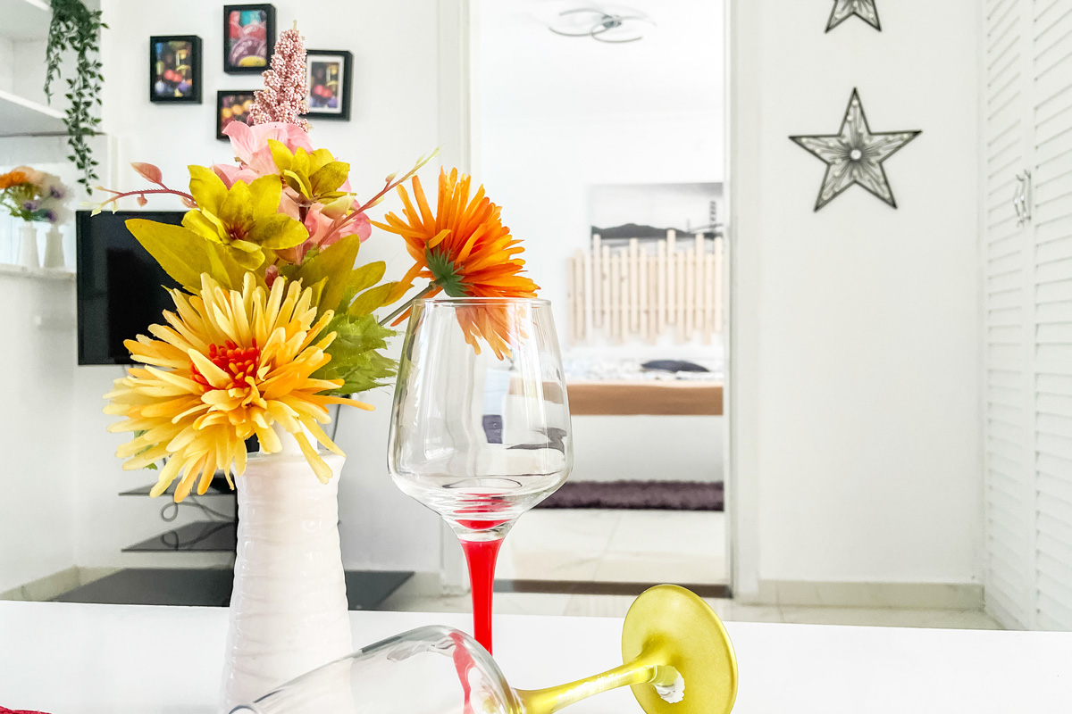 Elegant interior detail with a white vase of vibrant yellow and orange flowers, two decorative wine glasses, and a cozy bedroom visible through an open doorway in a bright, minimalistic setting.