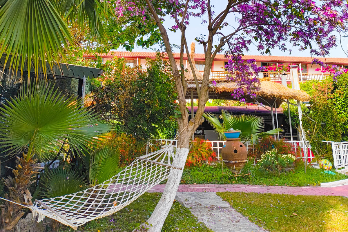 hammock-in-the-garden Cozy hammock tied between trees in a lush garden filled with palm leaves, vibrant flowers, and a large terracotta pot. Behind the garden is a traditional building with red rooftops and a shaded terrace, creating a peaceful Mediterranean retreat atmosphere.