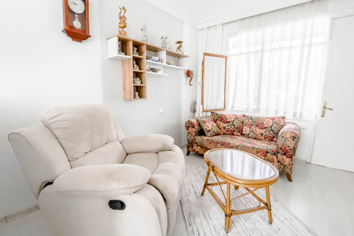 Cozy living room with a plush beige recliner, a floral-patterned sofa, a glass-top rattan coffee table, and wall-mounted shelves decorated with figurines, all softly lit by natural light through sheer curtains.