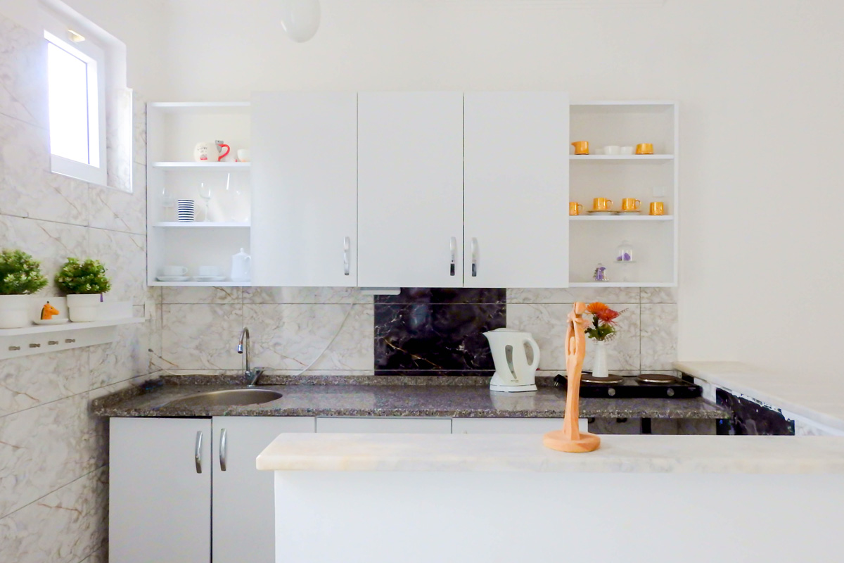 Bright kitchenette with white cabinets, marble-patterned backsplash, open shelves displaying cups and dishes, and a countertop with a kettle and decorative vase.