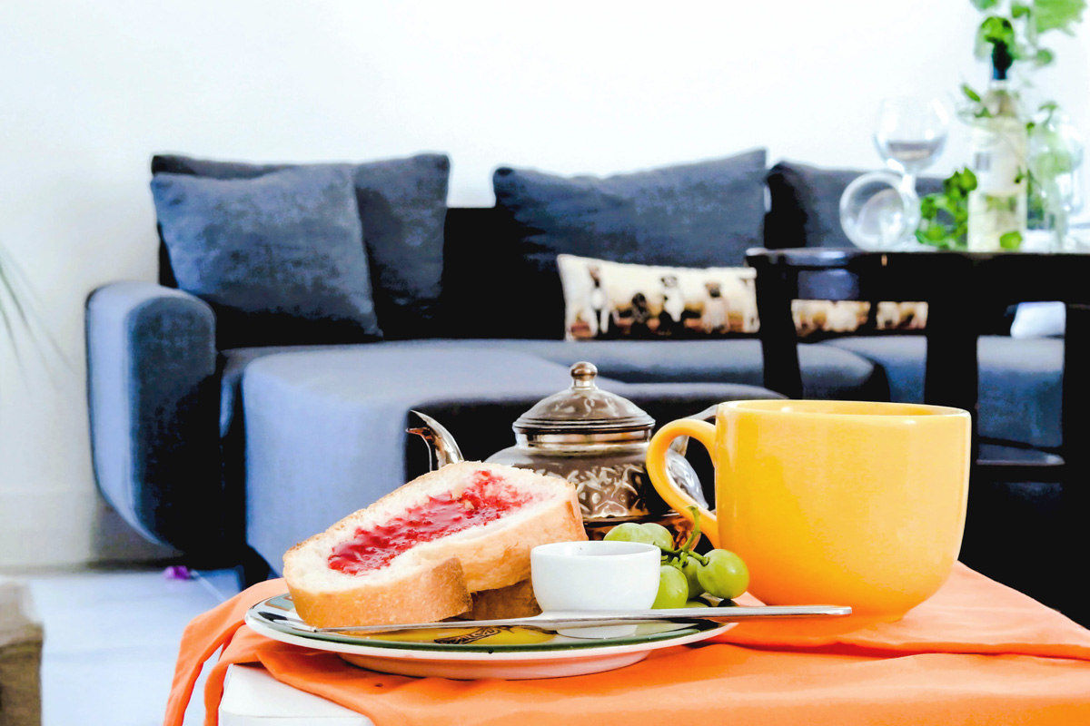 Cozy breakfast setup with toast and jam, green grapes, a yellow mug, and a decorative teapot on a white table with an orange cloth, set in front of a modern gray sofa with patterned cushions.