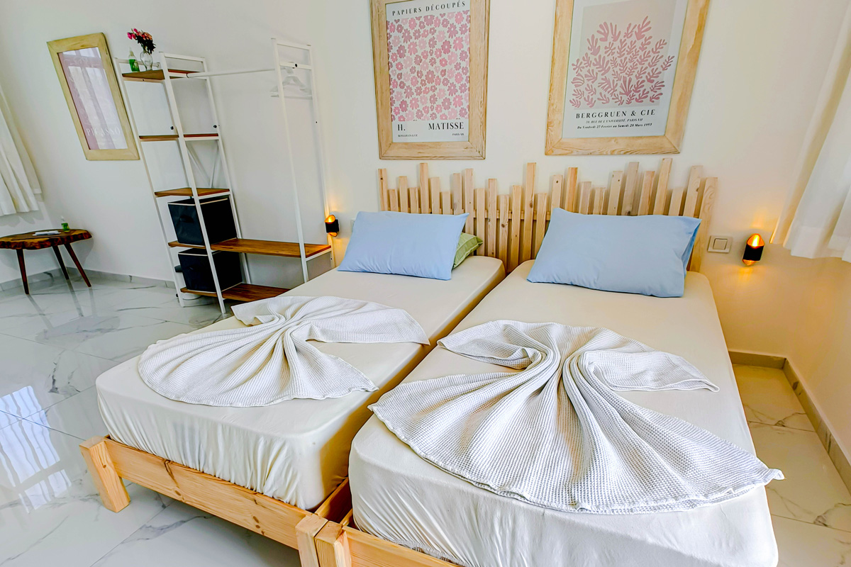 Modern guest room with two twin beds featuring light wood frames, blue pillows, and towels folded into decorative shapes, complemented by framed wall art and minimalist furniture.